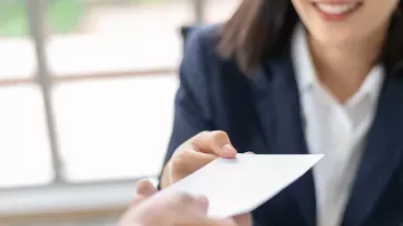 A woman in a suit hands over a white envelope with a smile.