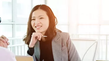 Woman in a gray blazer smiling during a conversation at an office desk.