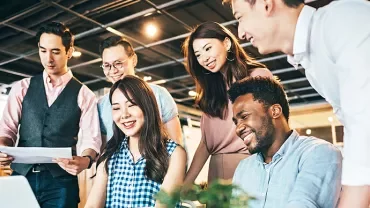 A group of six colleagues smiling and collaborating around a laptop in an office setting.