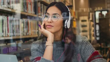 An East Asian woman in glasses looking out of a cafe, with headphones on.