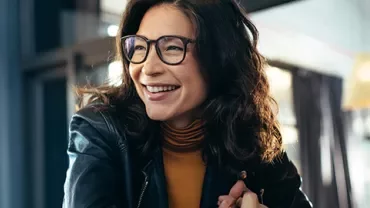 Woman in glasses and leather jacket smiling inside a cafe.