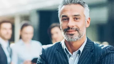 Smiling man in a suit standing confidently with blurred people in the background.