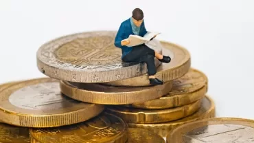 A male figurine sitting on a pile of gold coins reading a newspaper