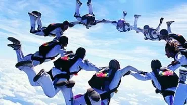 Group of skydivers forming a circle in the sky with clouds below.