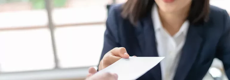 A woman in a suit hands over a white envelope with a smile.