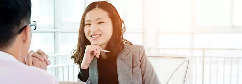 Woman in a gray blazer smiling during a conversation at an office desk.