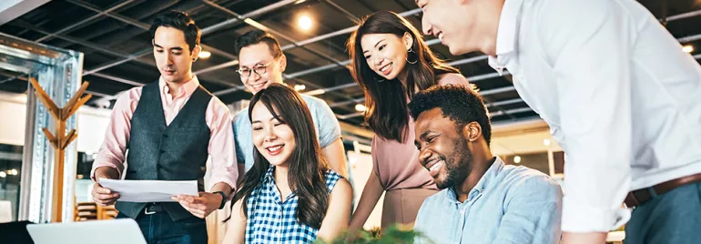 A group of six colleagues smiling and collaborating around a laptop in an office setting.