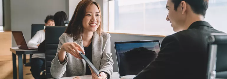 Two professionals having a conversation in an office setting with laptops.