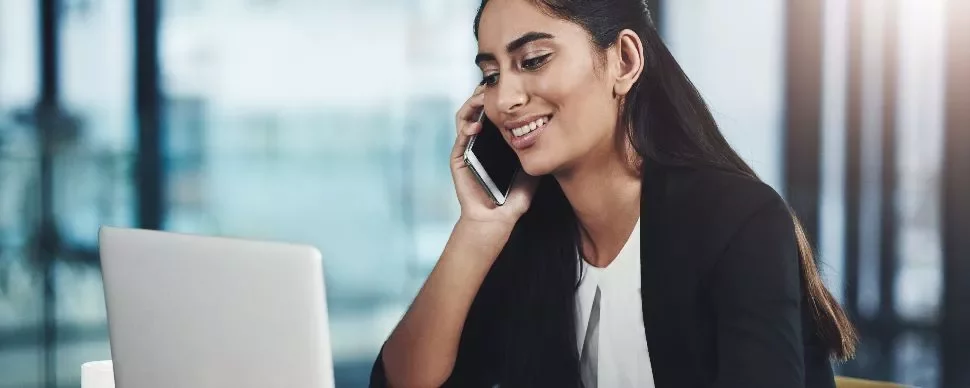 Woman talking on a mobile phone while using a laptop in an office setting.