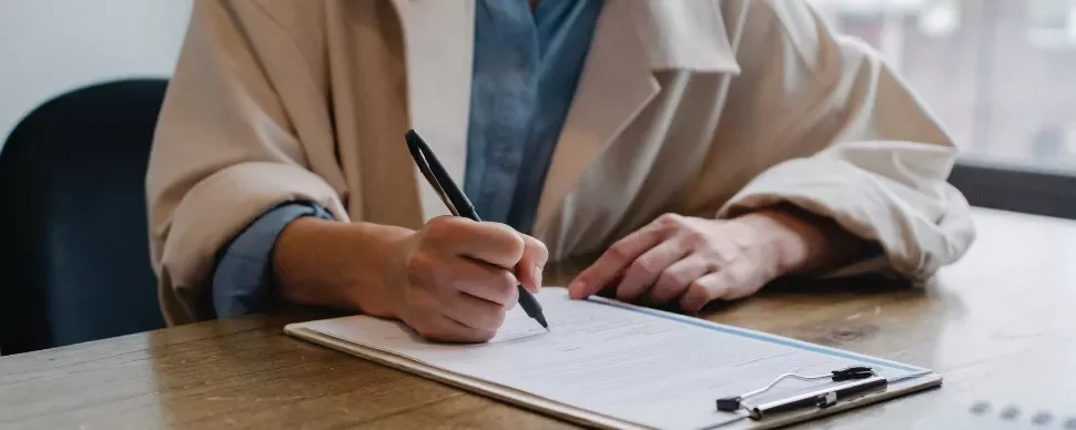 Person in a beige coat writing on a document at a wooden table.