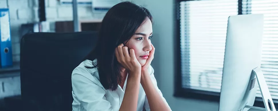 Woman sitting at a desk looking at her computer screen thoughtfully.