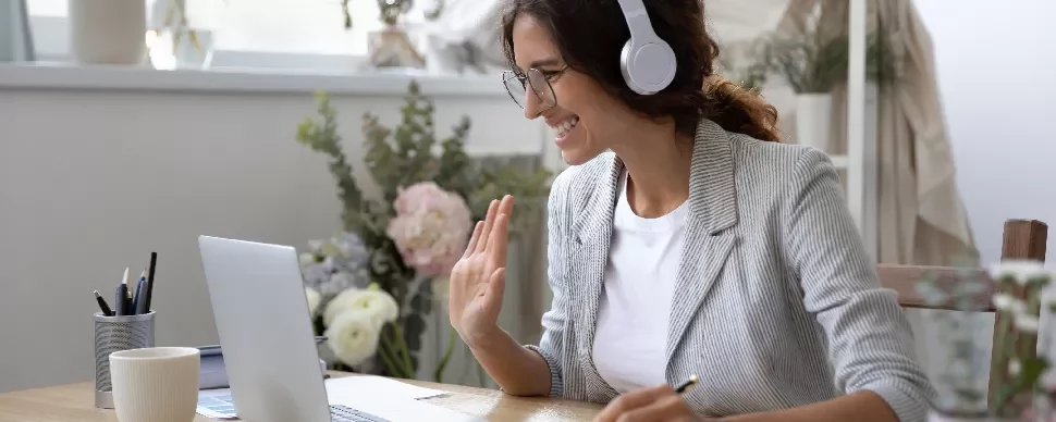 Smiling woman with headphones waving on video call at home.