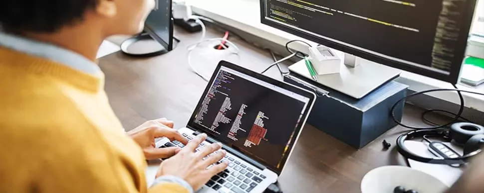 Person coding on a laptop with a large monitor in the background.