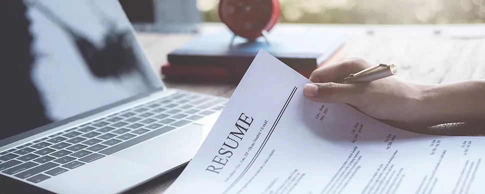 Person holding a resume in front of a laptop on a wooden desk.