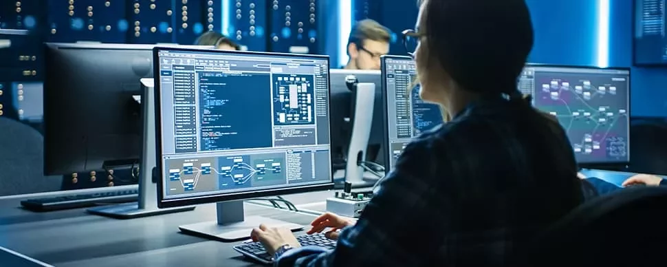 A person working at a computer with multiple screens displaying complex data and charts in a tech office.
