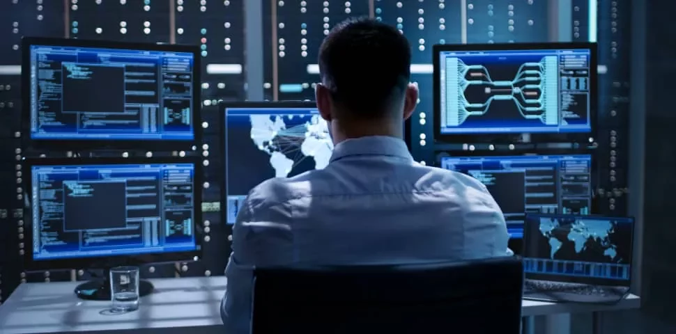 Man in front of multiple screens showing data in a dark server room.