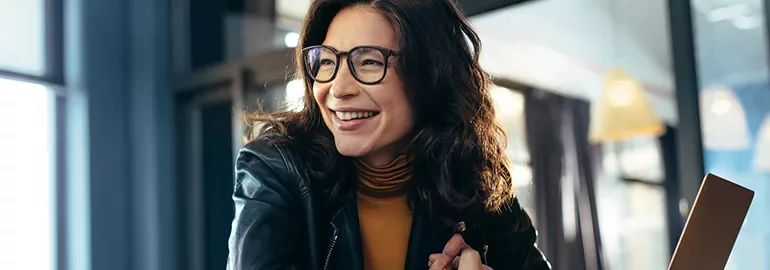 Woman in glasses and leather jacket smiling inside a cafe.