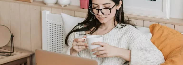 Woman with glasses holding a mug and using a laptop in a cozy room.